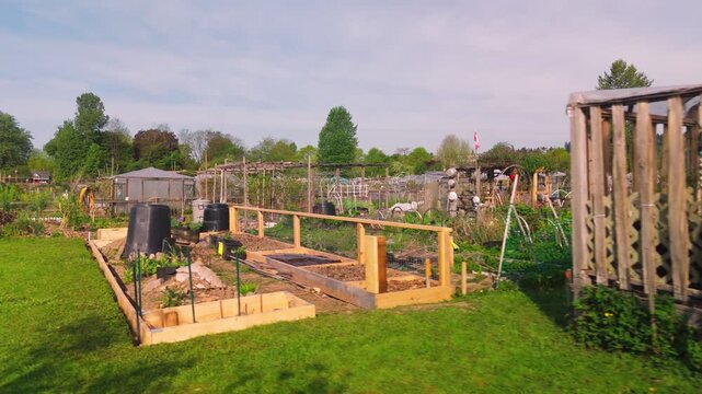 Panning perspective across a community garden allotment featuring newly constructed wooden raised beds and large black compost bins. The scene captures a variety of cultivation plots weathered