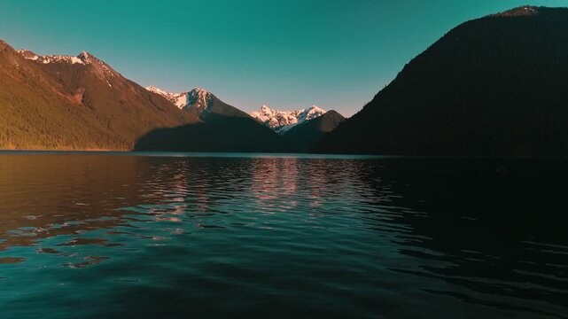 Expansive aerial view floating over dark calm lake waters stretching toward distant rugged alpine mountains topped with snow glowing brightly from setting sun approaching drone low altitude