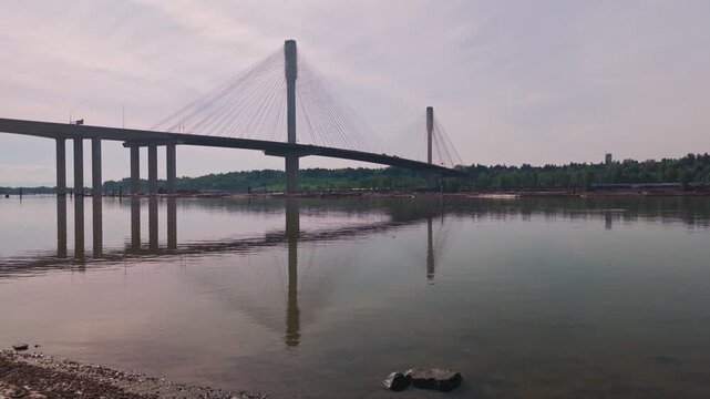 Cable-stayed bridge towers dominate the horizon as the camera glides over the gravel shoreline and glassy Fraser River water toward the massive architectural span approaching