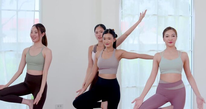 Asian adult female friends smile while holding tree pose, lifting arms for balance, core stability, posture control, concentration, flexibility and joyful wellness routine in bright studio class