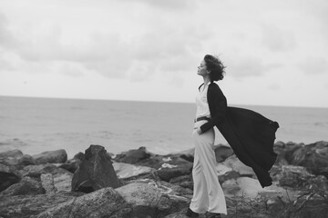 Naklejka premium Wind sweeps along the coast as a cape billows behind a figure on rugged rocks, gazing toward the horizon, capturing solitude, strength, and maritime escape in monochrome.