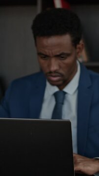 Vertical shot of black man sitting at desk, looking at laptop screen while working on it in his office