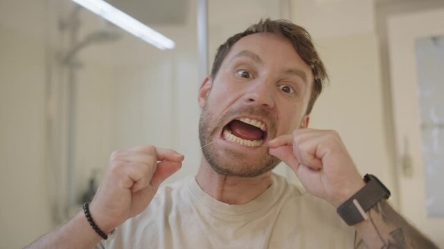 Young man with a tattoo making funny faces while flossing his teeth in front of a mirror