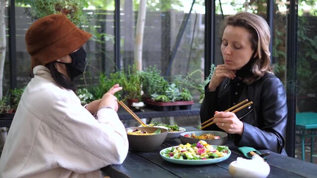 Two women in face masks sit at a restaurant table, briefly lowering masks to eat and chat, illustrating pandemic era dining, social distancing and cautious socializing