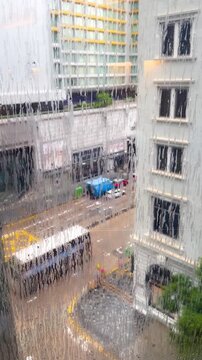 ฺBlurry video through a clear glass window shows rain falling outside in Hong Kong. Raindrops are drizzling down, tall buildings are visible, along with traffic, including buses and cars.