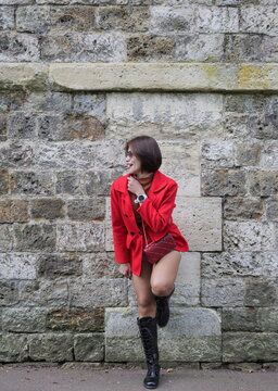 A woman in a red jacket and tall black boots leans against a stone wall with one leg raised