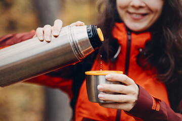 Fototapeta premium Smiling woman pours hot drink from metal thermos into cup outdoors. vacuum insulation technology, thermal efficiency engineering, functional product feature.