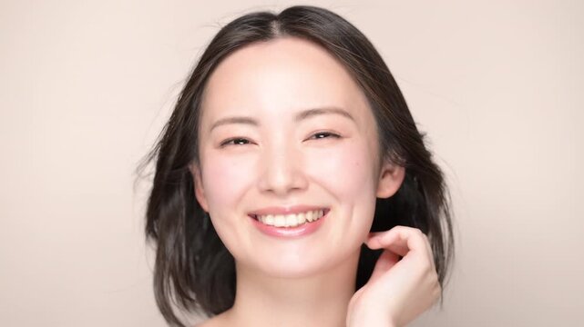 A woman with smooth, black hair blowing in the wind against a beige background, smiling with beautiful, clear skin&mdash;a close-up video showcasing skincare and beauty treatments, looking directly at the c