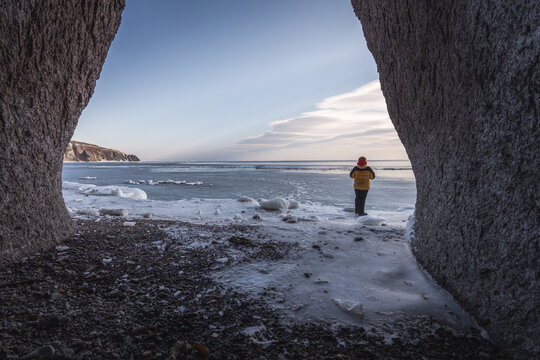 Person in yellow jacket standing on icy shore, framed by rock formations, overlooking frozen sea and distant cliffs under a clear sky during winter season