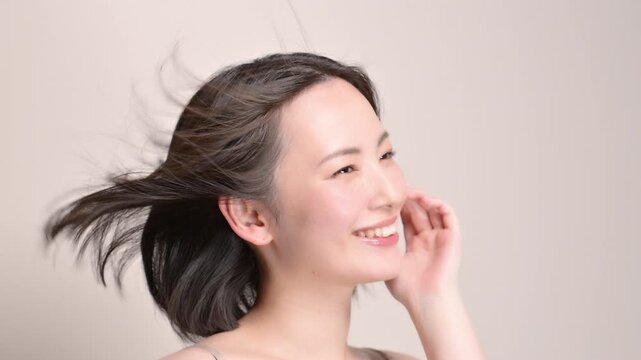 ベージュ背景の風になびくサラサラ黒髪と笑顔の女性美肌と透明感あるスキンケア美容やトリートメントイメージ頬に手で触れる動画　スローモーション右向き