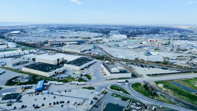 Industrial Corridor Logistics View. I-495 overpass intersects industrial zones near Christiana River. Dusty gravel mounds blend tan warehouse roofing, dark asphalt lines under blue sky, DE