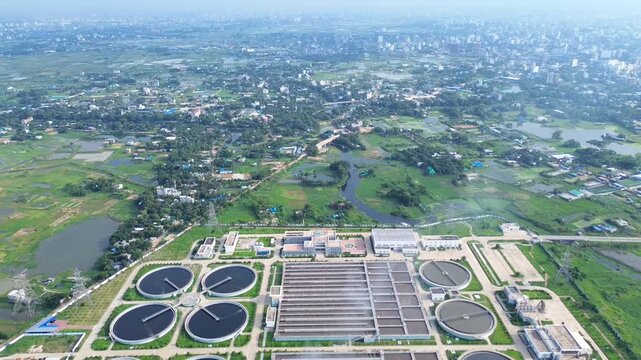 Aerial view of a large sewage treatment plant with circular clarifiers and rectangular tanks surrounded by green fields and urban landscape in Dhaka, Dhaka Division, Bangladesh.