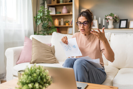 Young woman wearing glasses reviewing documents while using laptop at home, focused on paperwork, online study, remote job, planning tasks and freelance work with serious thoughtful expression indoors
