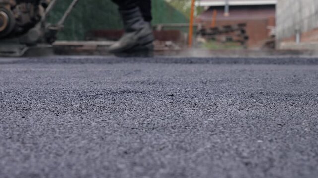 Fresh Asphalt Surface With Worker Using Vibratory Plate In Background
