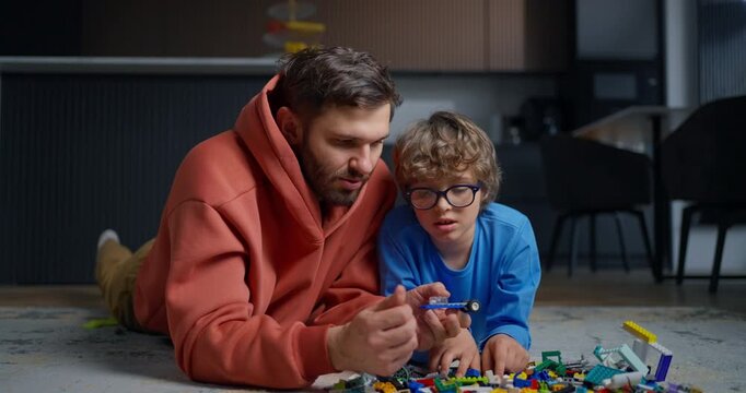 Father and young son lying on living room floor, bonding through creative construction play with colorful toy bricks in a cozy home interior