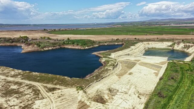 A large area shows several water bodies surrounded by sandy land and fields. There are patches of grass in the landscape. The scene is set under a cloudy blue sky.