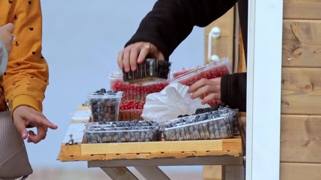 Close-up of hands arranging fresh berries in transparent containers at a wooden market stall. A customer in a yellow patterned jacket gestures nearby, creating a lively, authentic scene of everyday ma