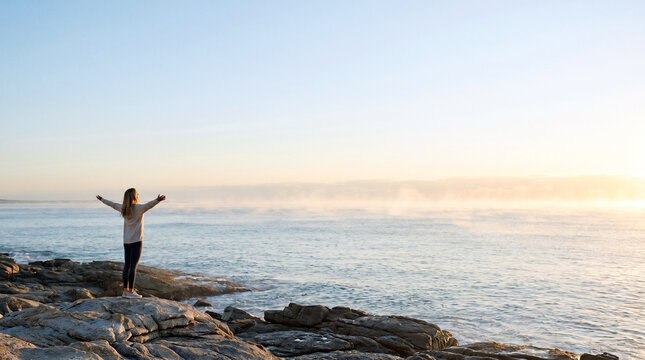 Young woman stands on a rugged rocky coastline with arms wide open, greeting a serene ocean covered in thick morning mist at sunrise. The warm golden light creates a peaceful, ethereal atmos