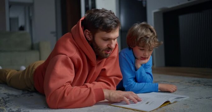 Father and young son lying on a living room rug, reading an open book together during a quiet family learning moment at home