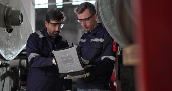 Two factory technicians review machine data on a laptop beside heavy industrial equipment in a traditional metal workshop, combining manual expertise, inspection, and modern diagnostics.
