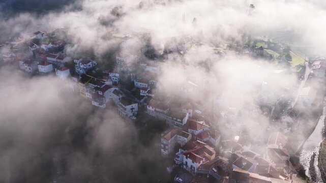 Aerial view of Avo village with red-tiled rooftops emerging from thick morning fog and mist along the Alva river in Oliveira do Hospital, Coimbra District, Portugal.