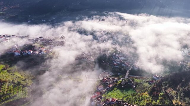 Aerial view of the historic village of Avo shrouded in thick white fog with green hills and traditional rooftops visible under soft sunlight in Oliveira do Hospital, Coimbra District, Portugal.
