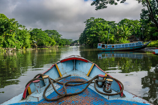 Boat travels on waterway in Barishal Bangladesh with green trees on the banks