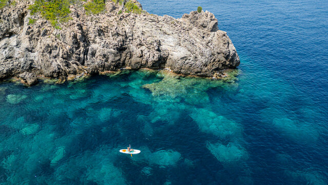 Tourist enjoys stand-up paddleboarding in turquoise waters near rocks in Mallorca, Spain, during a sunny day in summer