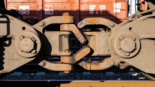 Close-up of a train coupling mechanism with rusted metal parts. Background features blurred freight train cars.