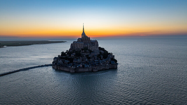 Aerial view reveals Mont St. Michel surrounded by water at sunset with cloudy sky in the background over France