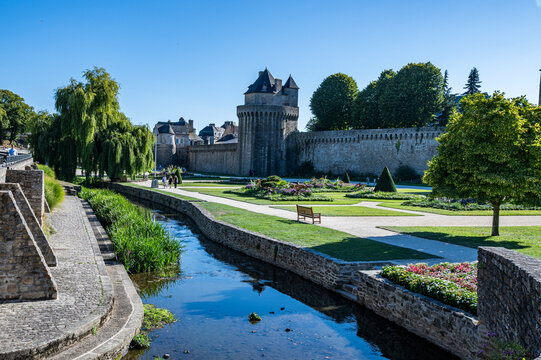 Visitors walk near the old stone walls and gardens of a historical site in France during a sunny day