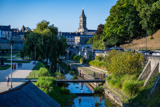 Visitors walk near the river and buildings on a sunny day in a French town with a historical clock tower and green trees