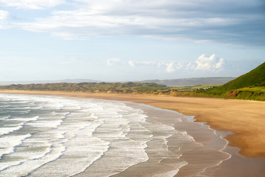 Evening light casts long shadows on Rhossili Bay Beach with gentle waves and sandy shores in Swansea, Wales