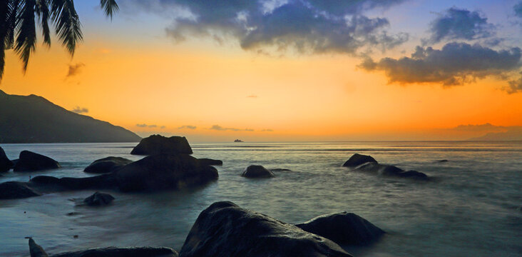 Sunset over the ocean at Beau Vallon beach in Mahe Seychelles with rocks and silhouettes of palm trees