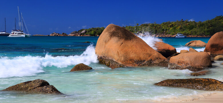 Waves crash against large rocks on the shore of Anse Georgette during a sunny day in Seychelles