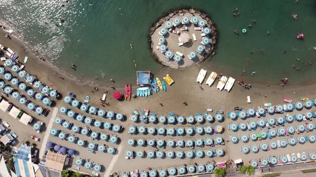 Aerial view of a crowded beach with blue umbrellas and a circular stone platform in the sea Imperia, Liguria, Italy.