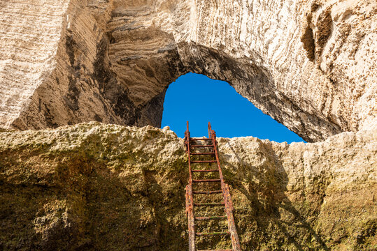 Rusty ladder leads up to natural arch with sunlight shining on chalk formations in Etretat, Normandy, France