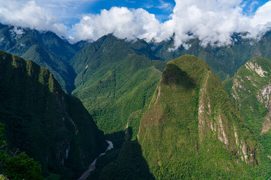 Mountains surrounding the Inca site at Machu Picchu display lush greenery and a distant river beneath a blue sky with clouds