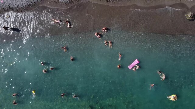 Aerial view of people swimming and relaxing in clear turquoise water along a dark sand beach with gentle waves in Imperia, Liguria, Italy.
