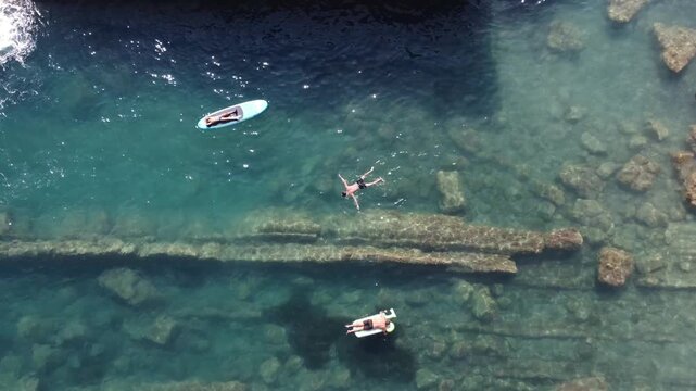 Aerial view of people swimming and paddleboarding in the crystal clear turquoise waters along the rocky coast of Imperia, Liguria, Italy.