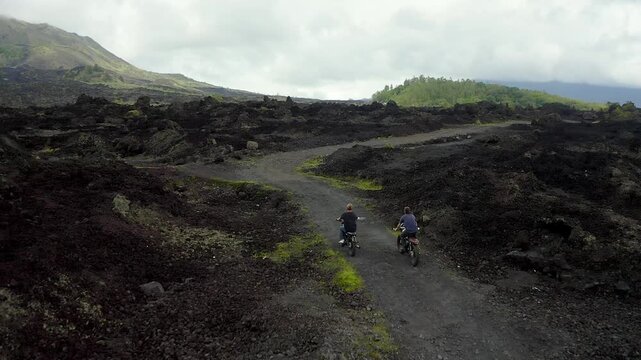 Aerial view of two motorcyclists riding on a winding dirt path through the dark volcanic lava fields of Batur volcano under a cloudy sky in Kecamatan Kintamani, Bali, Indonesia.