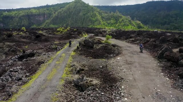 Aerial view of two people riding motorcycles on dirt paths across the black lava fields of Batur volcano with green hills in Kecamatan Kintamani, Bali, Indonesia.