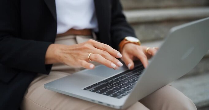Businesswoman typing on laptop keyboard while sitting outdoors on stairs.