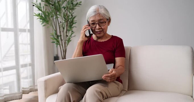 Senior asian woman sitting comfortably on a sofa at home, smiling while talking on a smartphone and using a laptop, effectively multitasking with modern technology