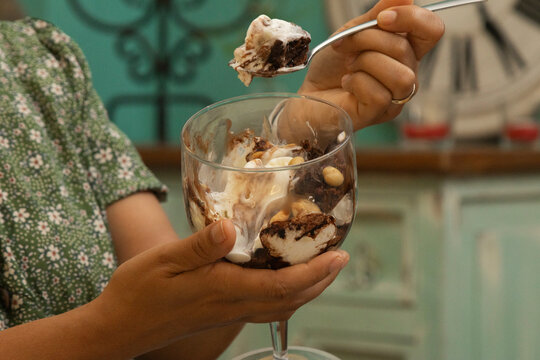 Person eating a decadent chocolate ice cream sundae with brownie pieces and nuts from a glass dish