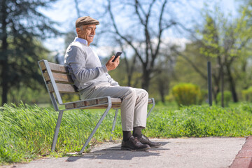 Fototapeta premium Elderly man sitting on a park bench with a smartphone