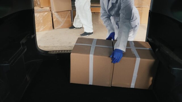 Medical workers in protective suits loading medicine cardboard boxes into car trunk for distribution.