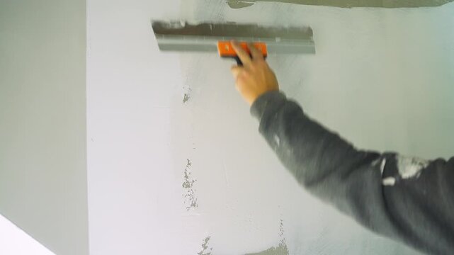 Worker uses spatula to level surface. Acrylic putty. Finishing work at construction site. Process of leveling and filling wall. Hand with trowel applies filler to concrete wall. Heavy manual labor.