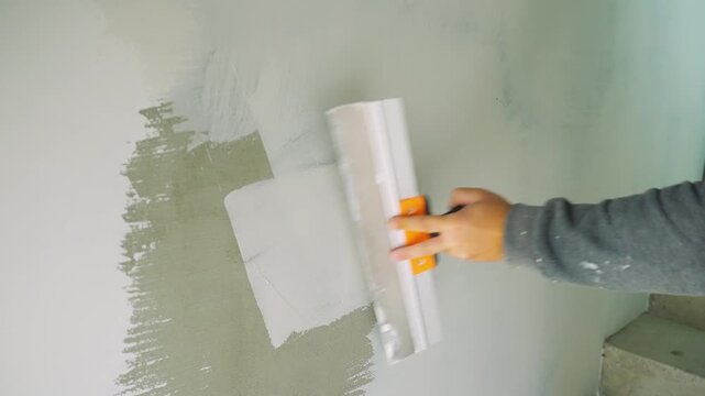 Leveling, filling concrete wall with acrylic finishing putty. Handyman applies putty to concrete wall using trowel, using left-to-right strokes. Heavy manual labor. Trowel is used to level surface.