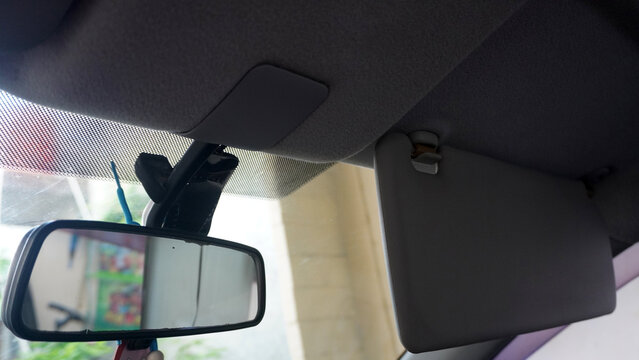 Interior view of a car showing the roof area above the windshield. A rearview mirror reflects part of the outside scene, while one sun visor is folded up and another down.